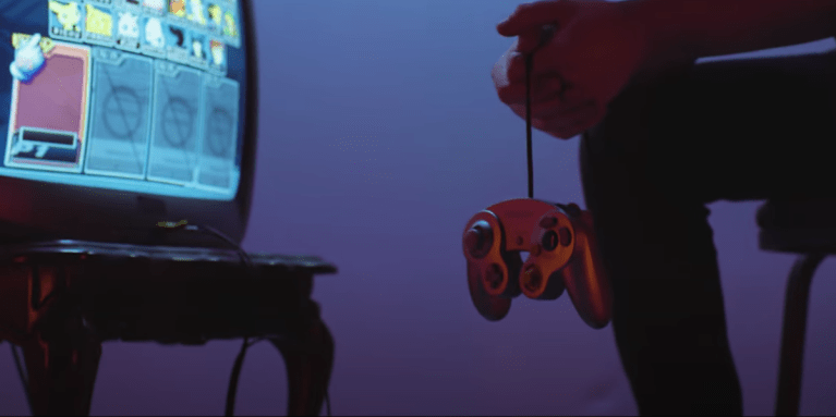 A player dangles a Gamecube controller from their hand in front of a tv showing the Melee character select screen.