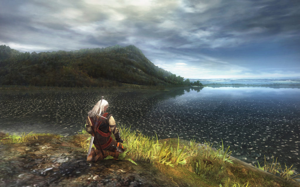 A screengrab from The Witcher game showing Geralt kneeling in front of a large body of water with a mountain in the distance