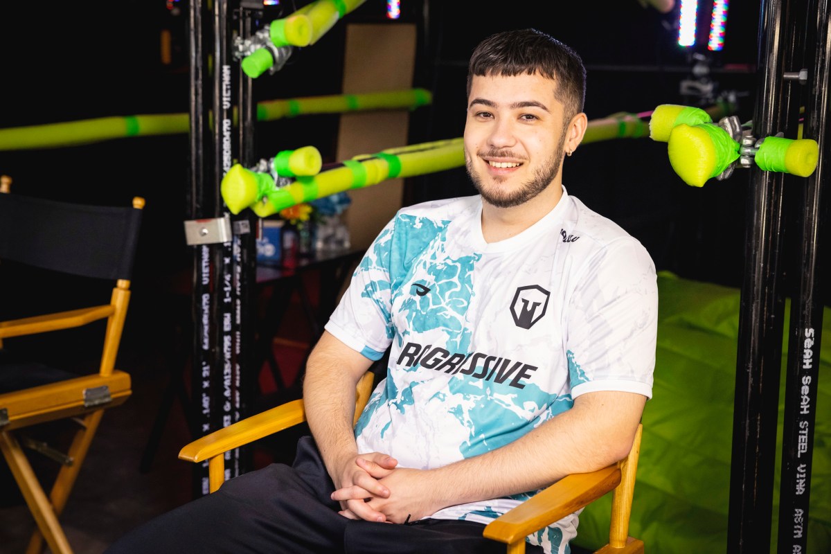 Mid laner Bolulu sits in a chair in a white Immortals jersey with various blue spots on it, while he smiles at the camera.