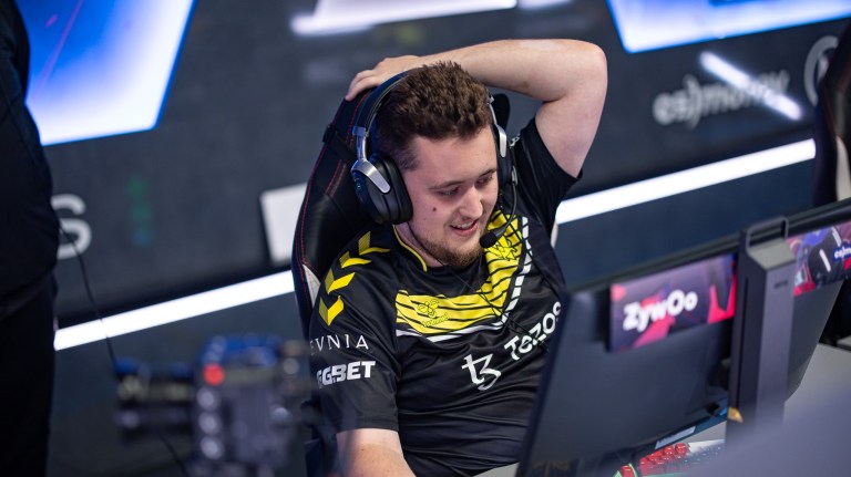 Photo of ZywOo, one of the best CS:GO players in the world. He's a white, has short hair, and is sit on the chair in front of gaming monitor with one hand behind his head and the other hand is holding the mouse.