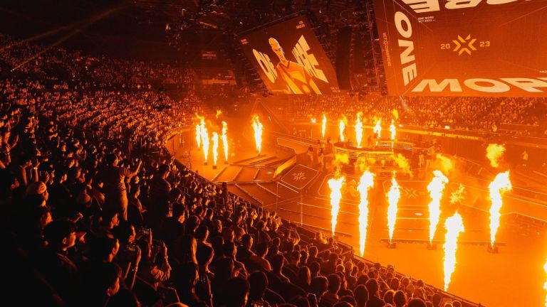 A massive crowd watches large screens that read "ONE MORE". Their silhouettes are lit up by multiple columns of fire.