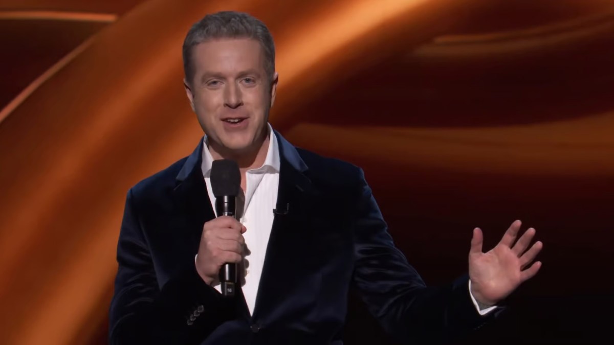 geoff keighley wearing a black suit jacket and white shirt, holding a microphone in his right hand with his left extended