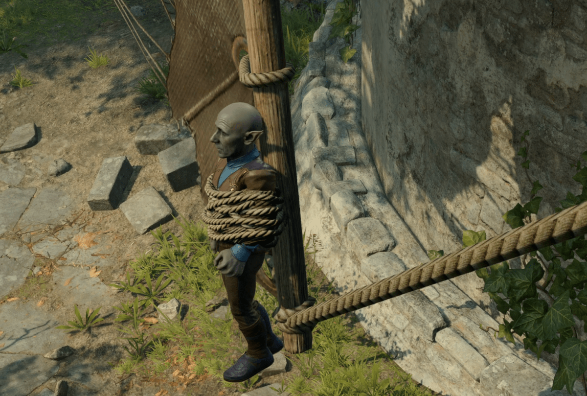 A small, grey-skinned gnome is tied to a windmill with rope.