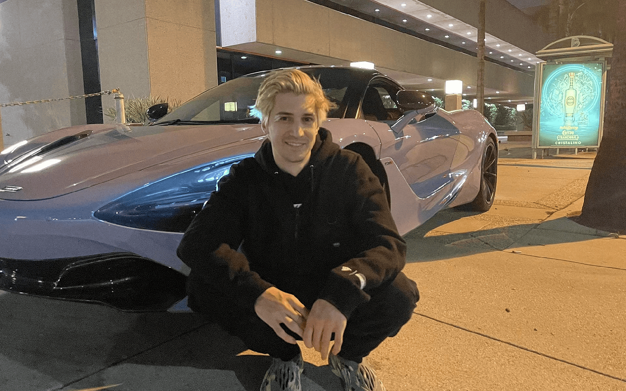 There is a picture of xQc sitting in front of his car. The car is white, and there is a blue sign in the background. 