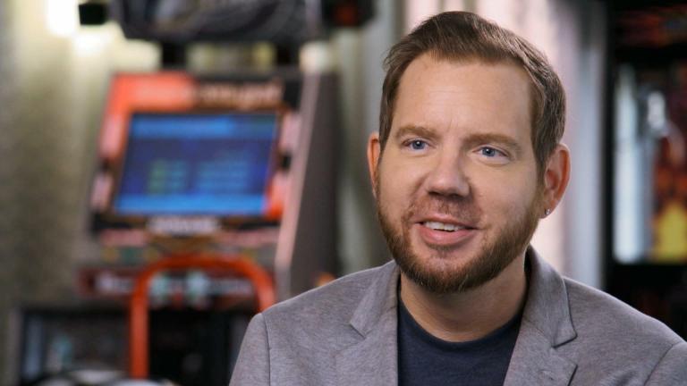 A bearded man with blue eyes and neat, brunette hair sits in front of a chair and arcade machine.