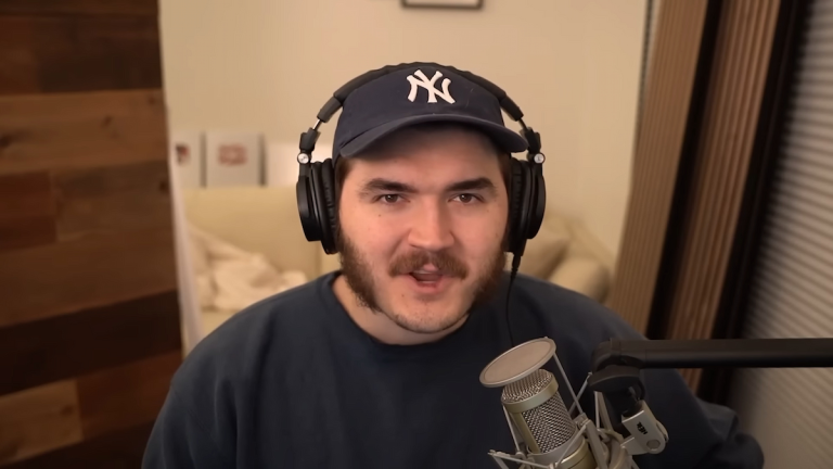 A man with mutton chops and a baseball cap wears earphones while addressing the audience through a silver mic.