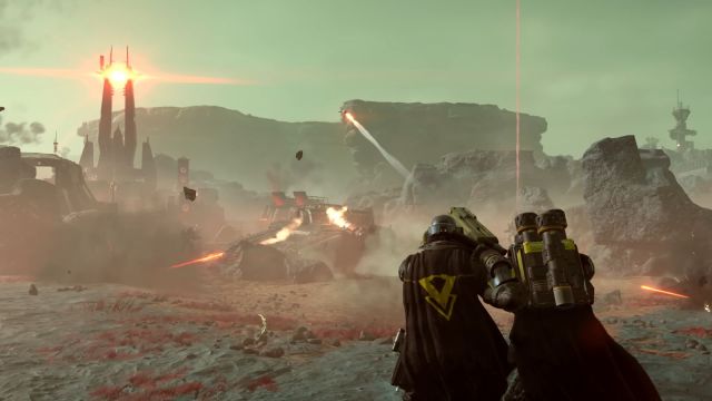 Image of Helldiver's 2 players firing a rocket launcher toward the sky. There is a barren planet backdrop in front of them and the sky is a dark to light green gradient.