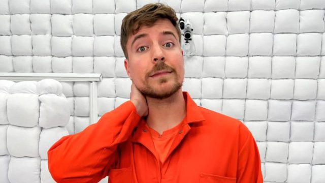 MrBeast, standing in a white padded cell, with his right hand on his neck, wearing an orange jumpsuit.