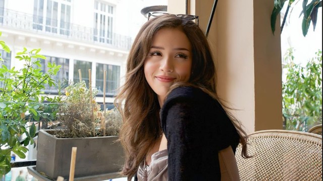 Pokimane sitting at a table, with sunglasses on her head. A yellow wall is behind her, with plants in the background.