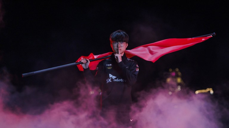 Faker, a League of Legends player, stands holding a finger to his mouth while holding a red T1 flag behind his back as smoke billows around him.