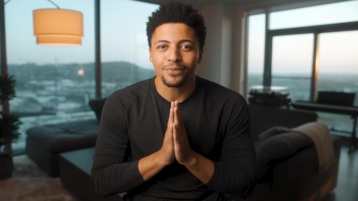 Content creator Myth holds his hands in a prayer stance while wearing a black shirt and speaking to the camera in his high-rise living room.