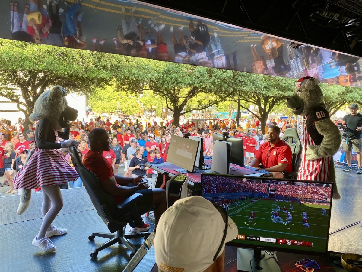 A group of people, including two in dog costumes, stand behind two men at computers on stage in front of a crowd.