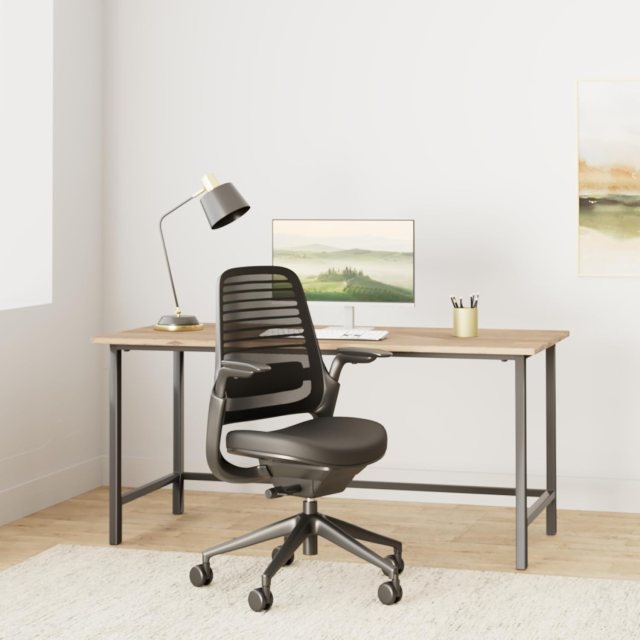 The Steelcase Series One in a brightly lit home office. The Series One is a gray rolling mesh chair with lumbar support, a cushioned swivel seat with foam filling, and wheels. More details below.