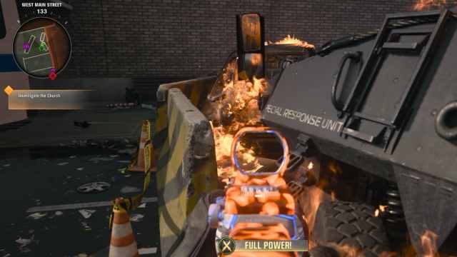 A player looking between a barricade and a destroyed tank towards a burnt-out car.