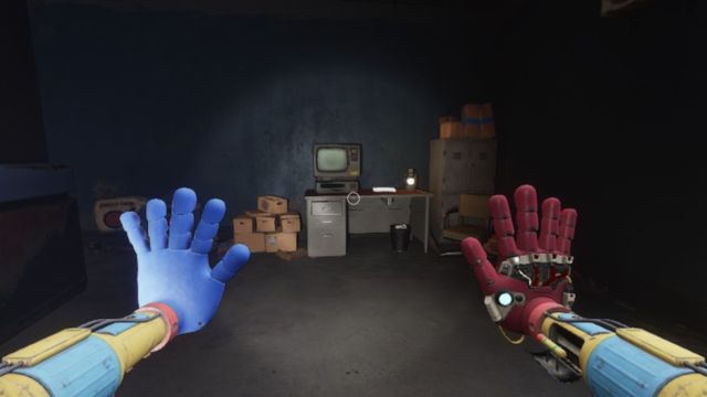 Looking at a VHS player and tape on a computer desk with a cabinet to the right and a pile of cardboard boxes to the left
