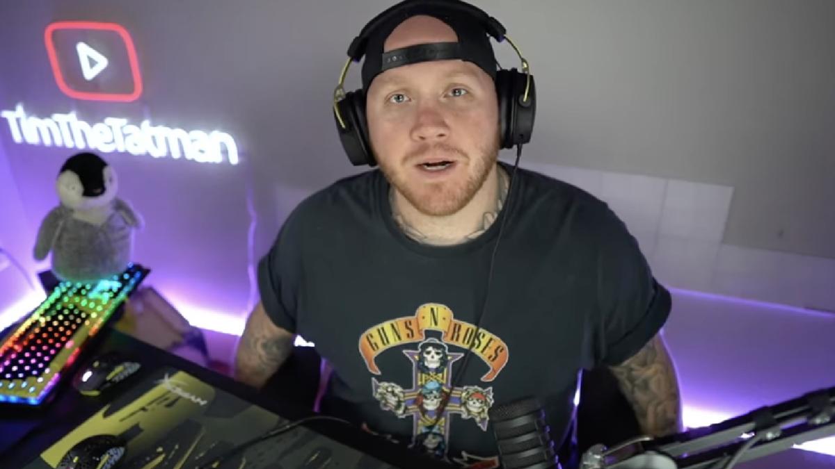 A man in a backwards hat and t-shirt with headphones on talks to the camera.