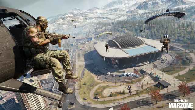 A soldier sitting at the door of a small helicopter while numerous other soldiers parachute around him above a stadium in Warzone.